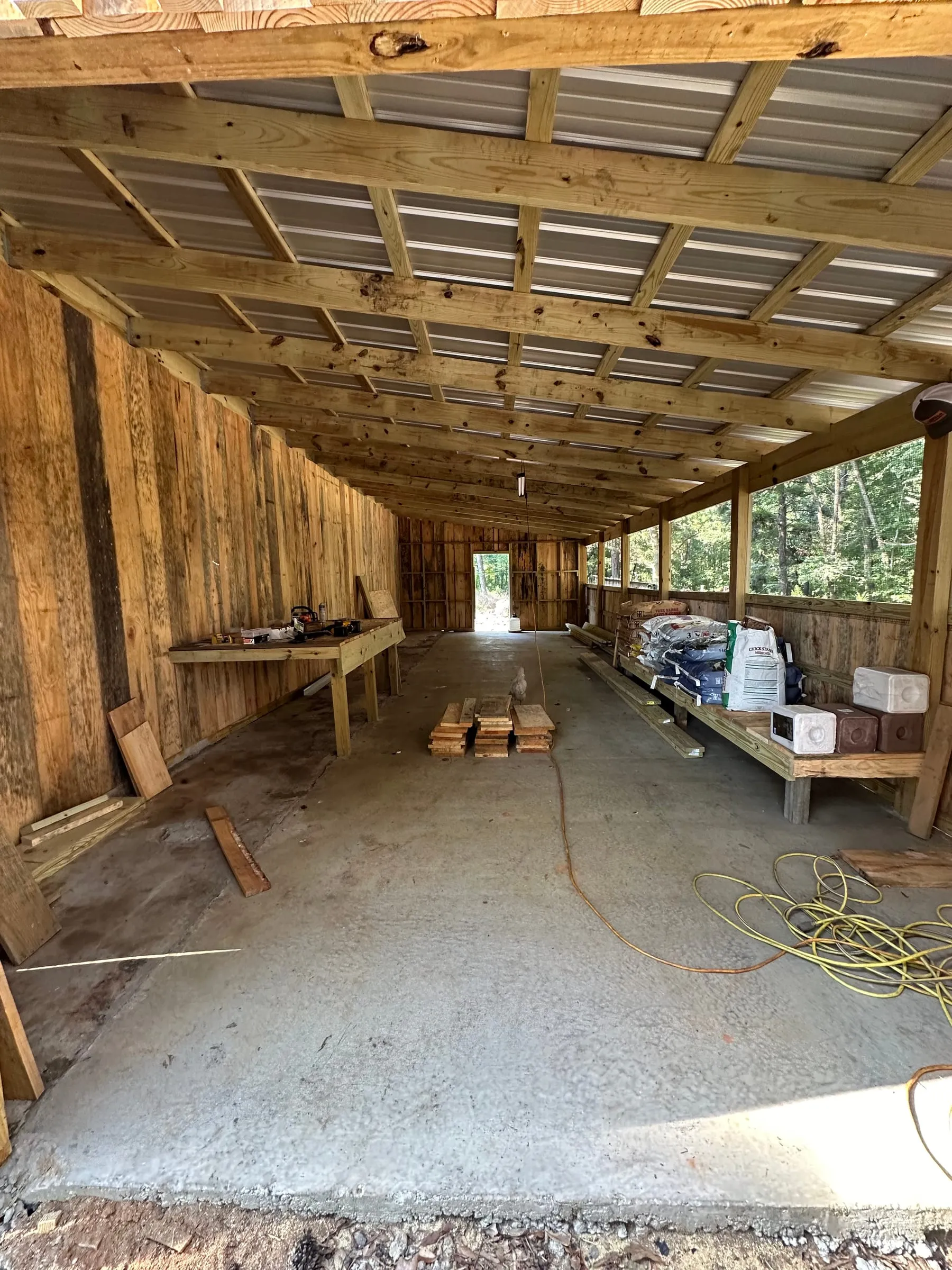 Inside the pole barn, board walls and exposed purlins over a concrete slab, with a built-in workbench and storage benches along an open lean-to bay.